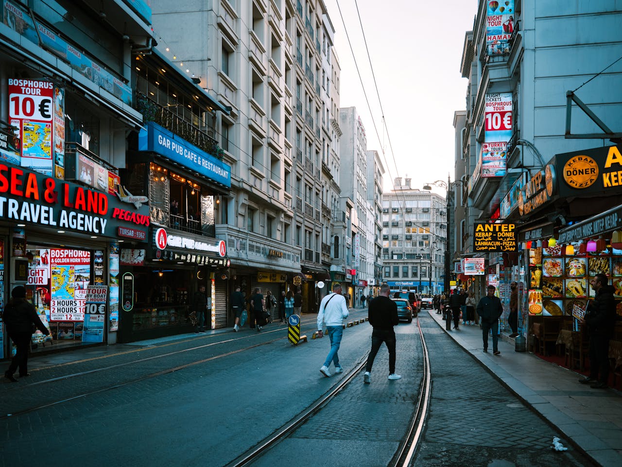 gallery-1 Vibrant street scene in Istanbul featuring shops, people, and tram tracks under a clear sky.