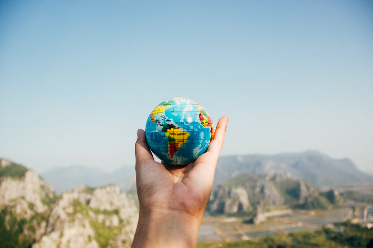 hero-img-01 Hand holding a globe against a mountain background symbolizing travel and exploration.
